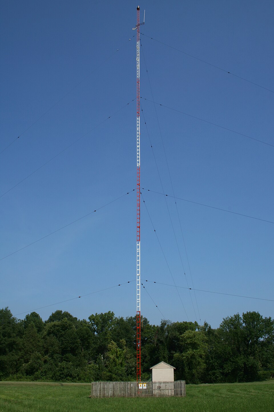A real red-and-white guyed lattice mast standing to full height against a clear blue sky, guy wires radiating down to ground anchors, equipment cabin at the fenced base