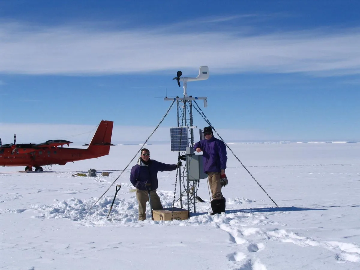 Two field engineers beside a guyed automatic weather station on an Antarctic ice sheet, a survey aircraft behind them